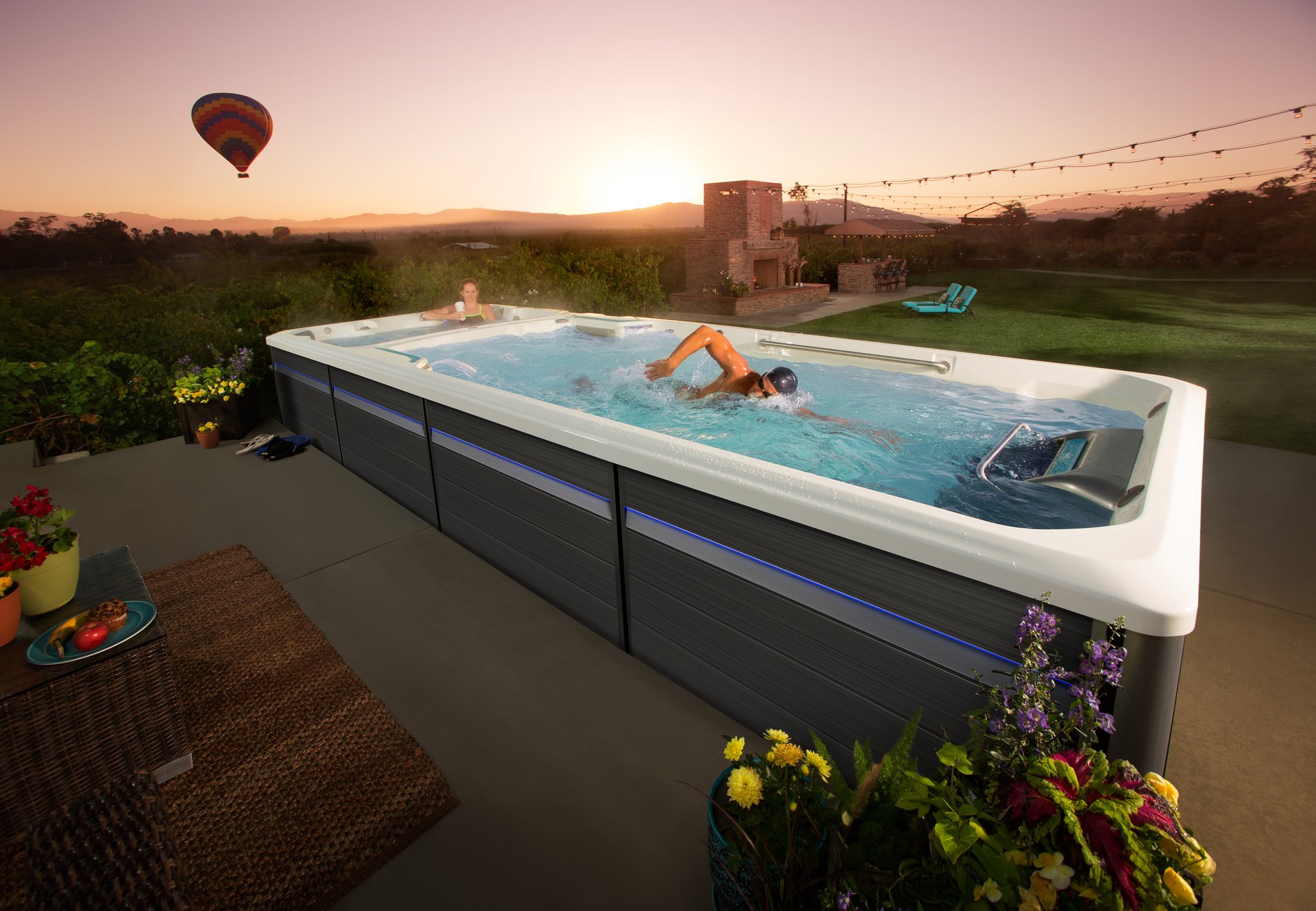 An Endless Pools swim spa on a patio at sunset, someone swimming against the current as a hot-air balloon drifts over the hills