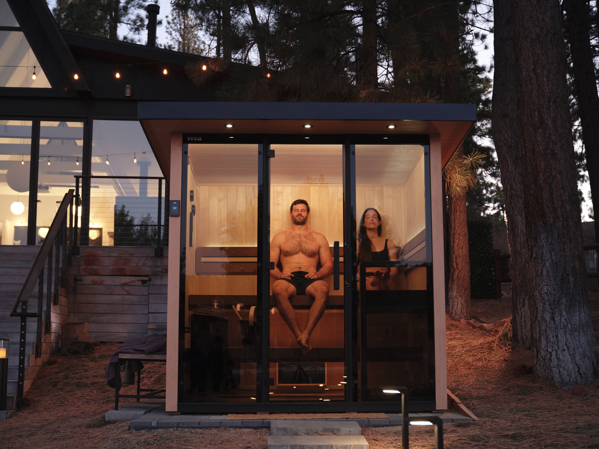 A glass-front Tylo Lulea sauna glowing warm at dusk in a forest clearing